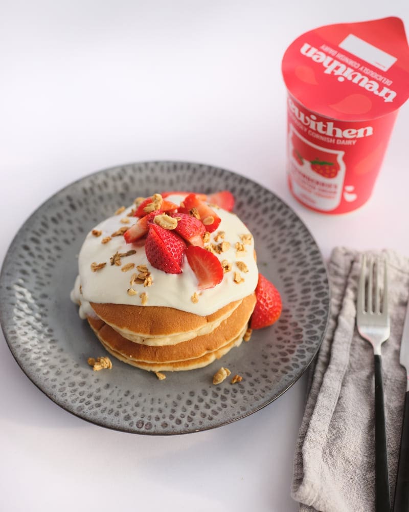 Pancakes with strawberry yoghurt and strawberries next to a tub of trewithen strawberries and clotted cream yoghurt