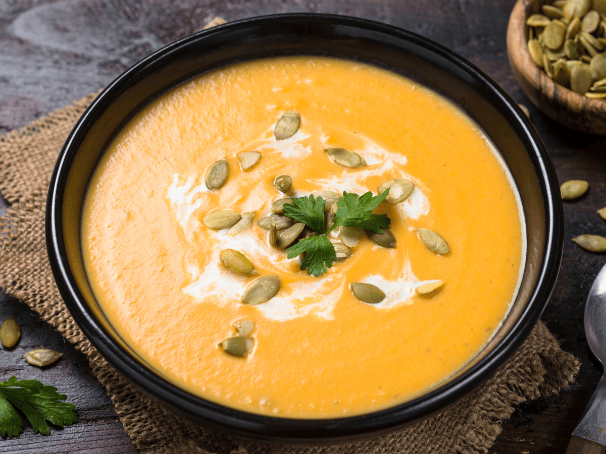 Pumpkin And Carrot Soup displayed in a black bowl topped with some yoghurt, herbs and seeds